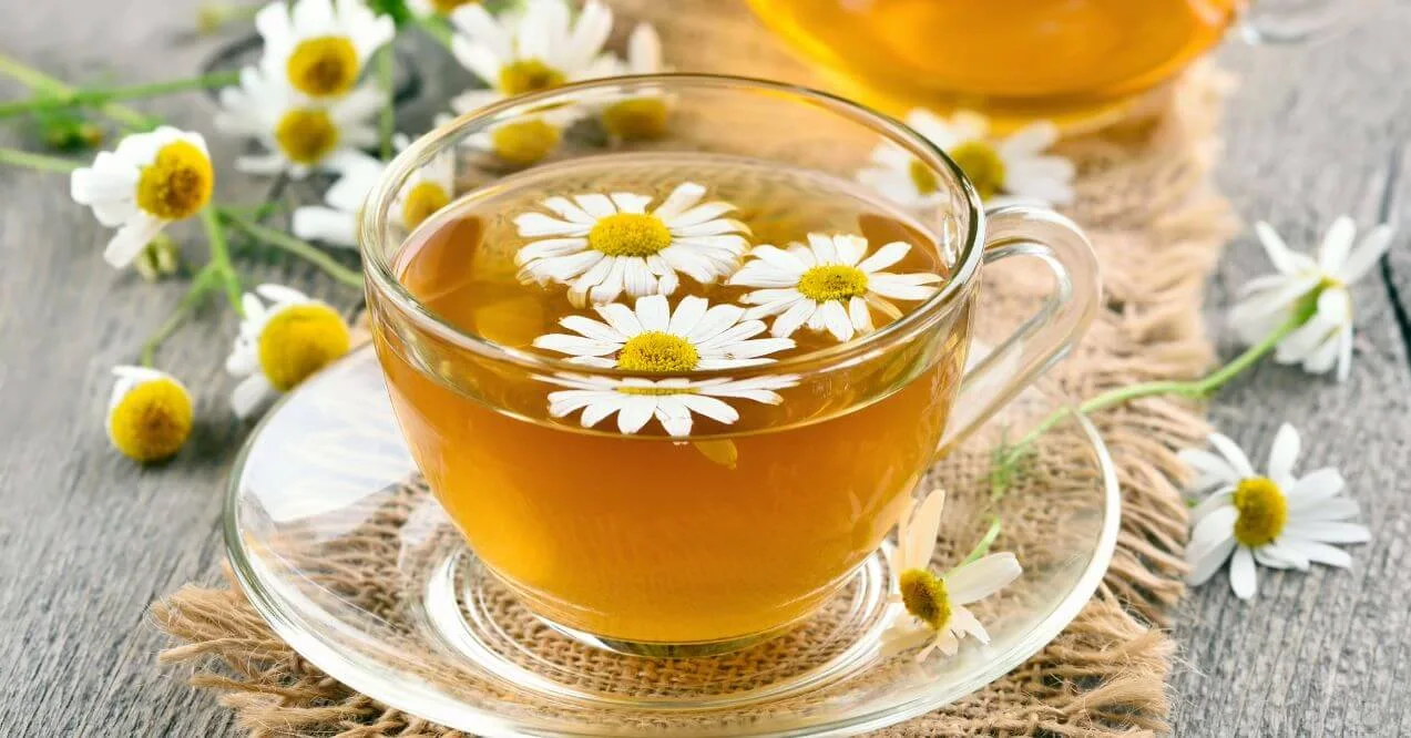 A glass cup of chamomile tea with floating chamomile flowers on a wooden surface.