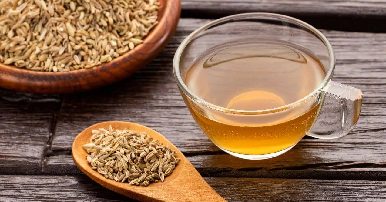 A glass cup of fennel tea with fennel seeds in a wooden spoon and bowl on a rustic table.