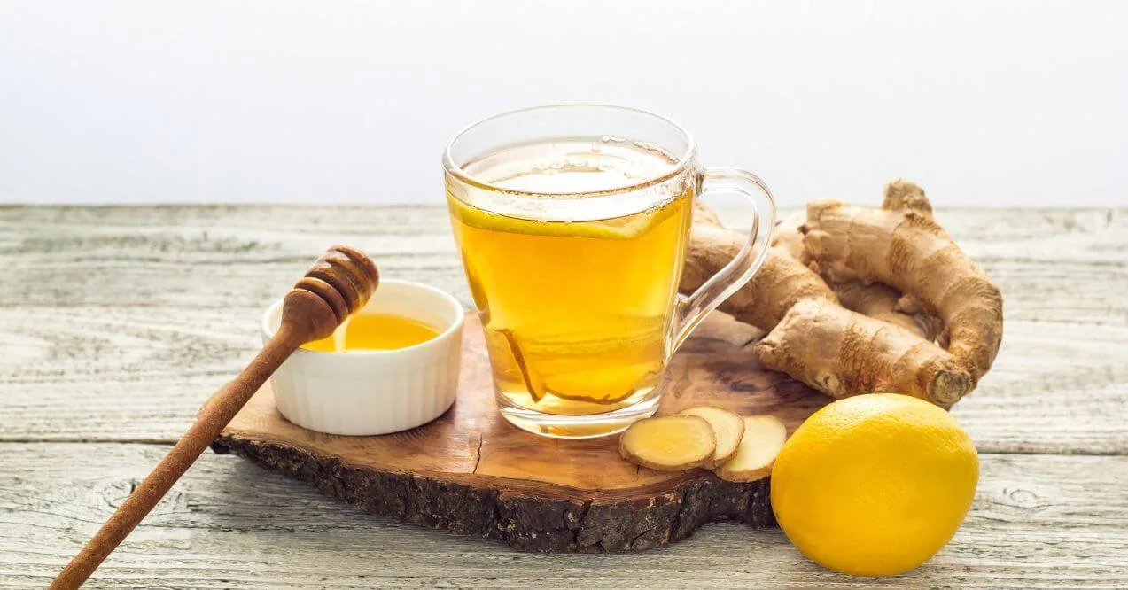 A glass mug of ginger tea with fresh ginger slices, lemon, and honey on a wooden board.