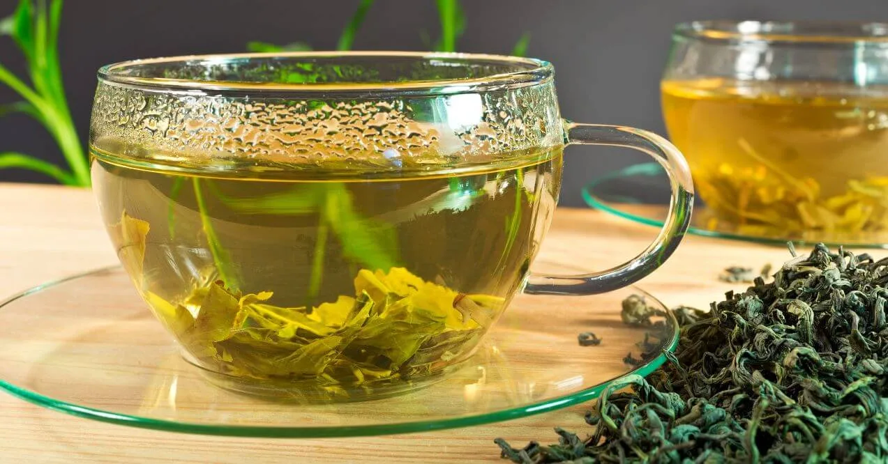 A glass cup of green tea with loose leaves and steam rising from the surface.