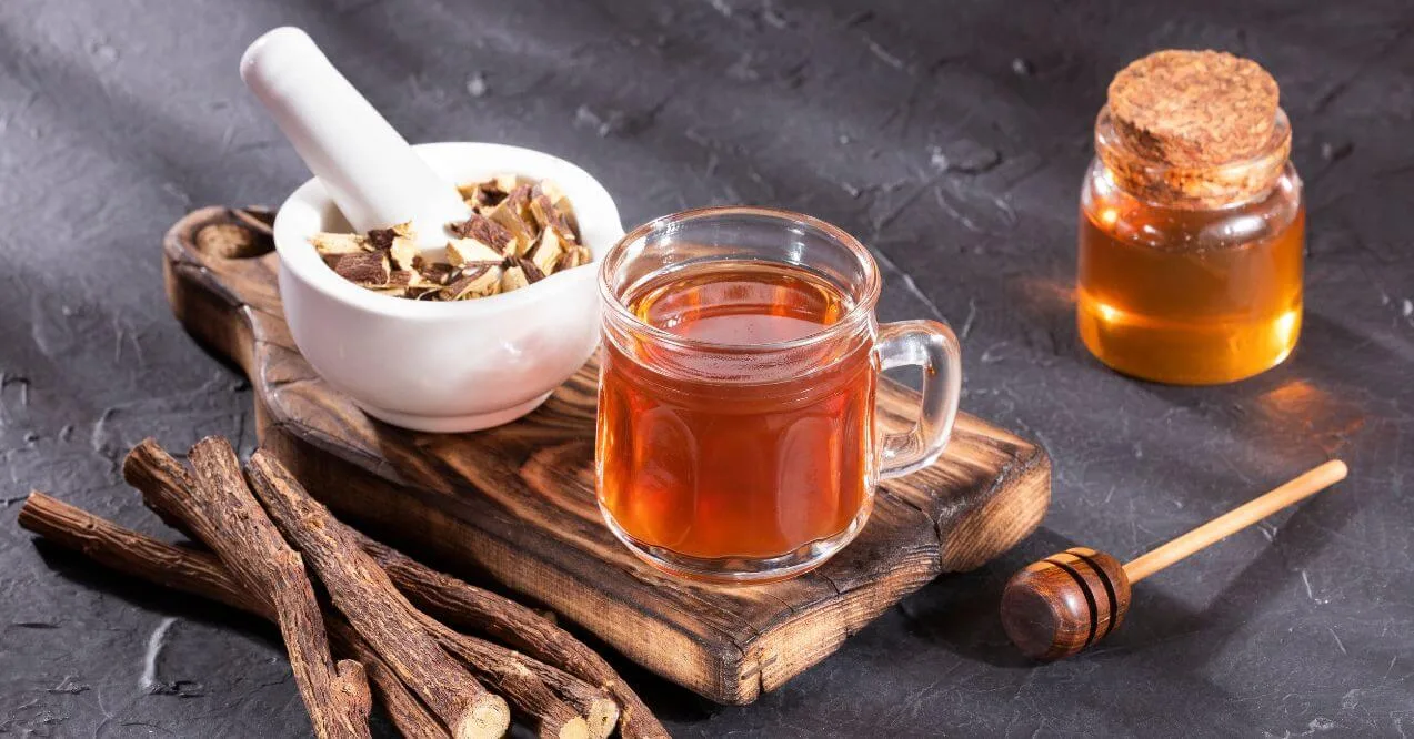 A glass mug of licorice root tea with dried licorice roots, honey, and a mortar and pestle.