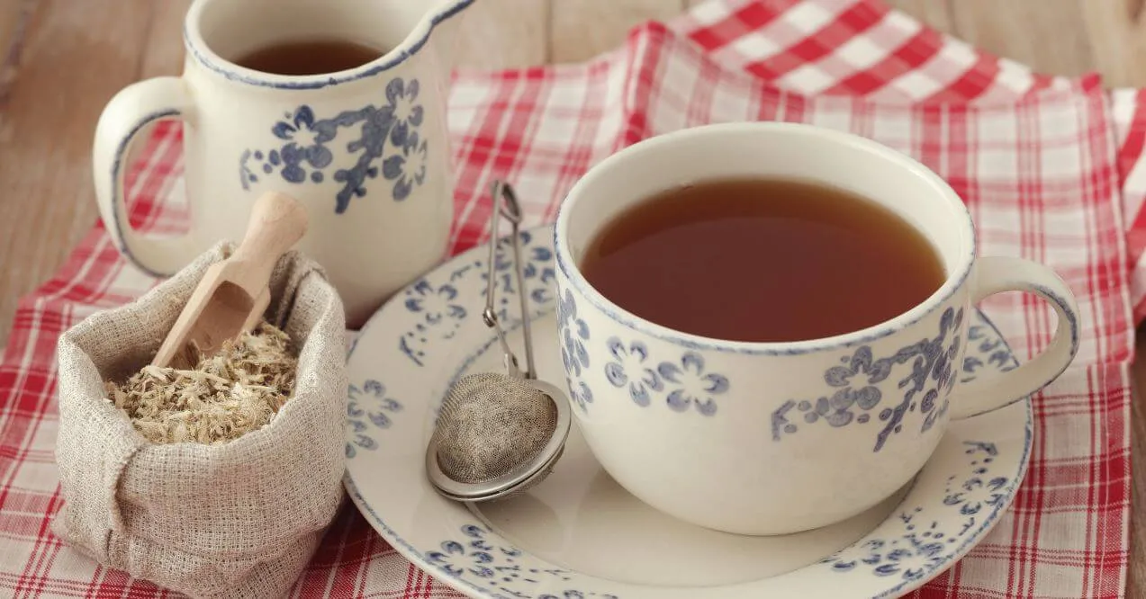 A blue and white teacup with marshmallow root tea, a sack of dried herbs, and a tea strainer.