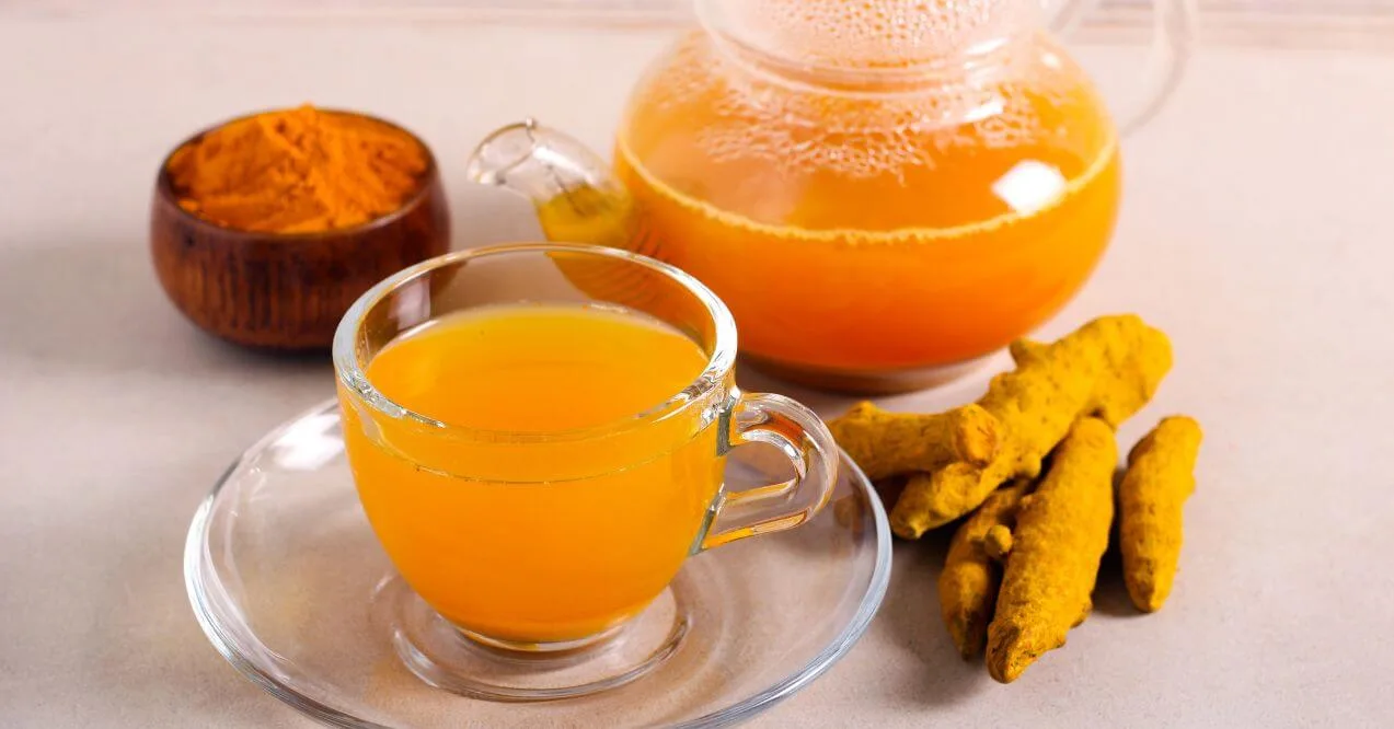 A glass cup of bright yellow turmeric tea with fresh turmeric root and powder.