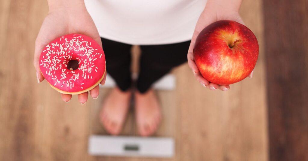 Woman Measuring Body Weight on Weighing Scale Holding a Donut and an Apple