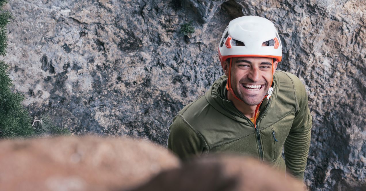 A man wearing a helmet and a green jacket is smiling at the camera. He is standing on a rocky surface, possibly a mountain or a cliff.