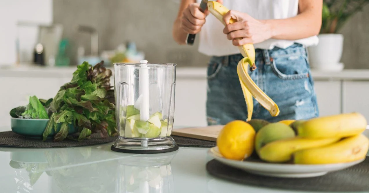 Young slim woman in white t-shirt and blue jeans cooking smoothie with bananas healthy food in kitchen at home.