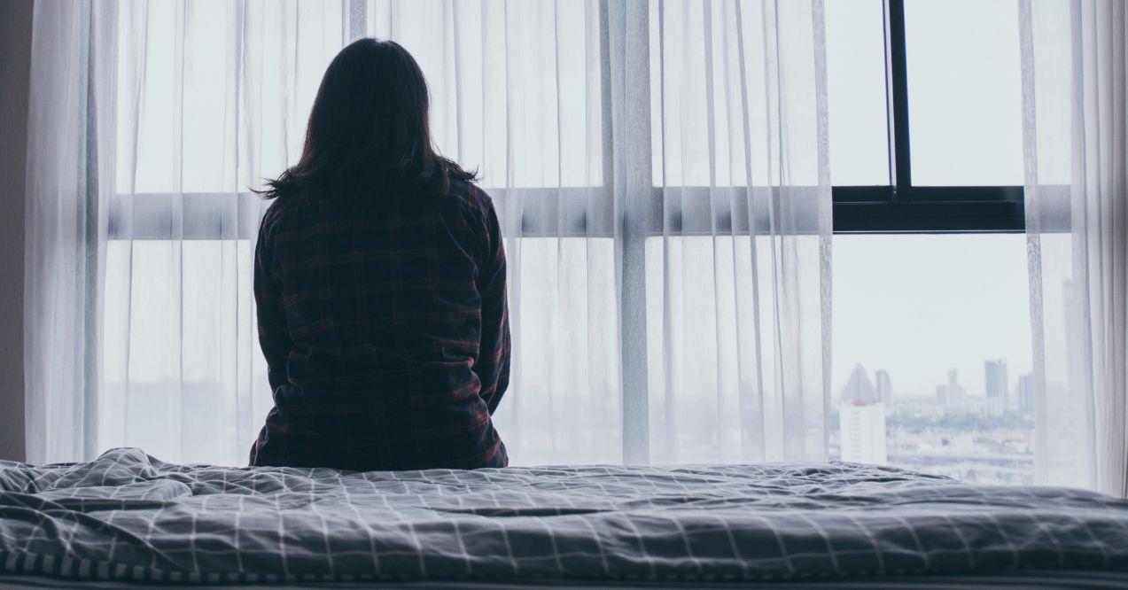 A woman sitting on a bed looking through the window in a dark room. Anxiety and depression.