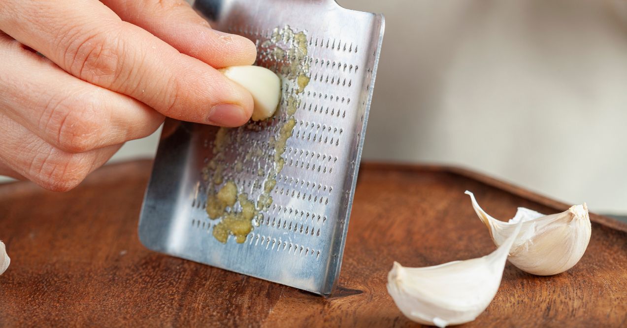 Hands of a caucasian woman grating garlic cloves using shovel shaped metal mini grater on a wooden plate.