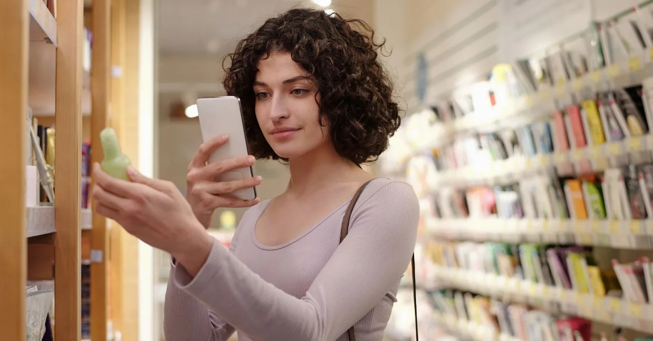 Young female consumer with dark curly hair taking picture or scanning qr code of skincare product in plastic bottle in cosmetic supermarket