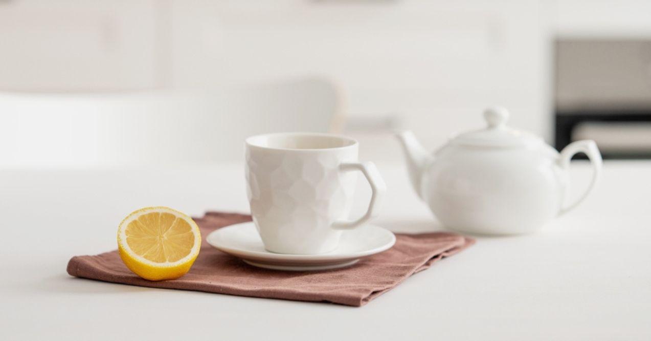A cup of tea with lemon and a teapot on a white table. Kitchen with wooden empty countertop and brick wall in the background.