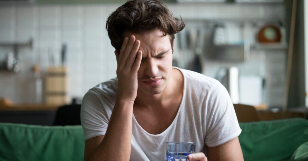 a man is holding one hand to his head due to a headache while holding a glass of water in his other hand