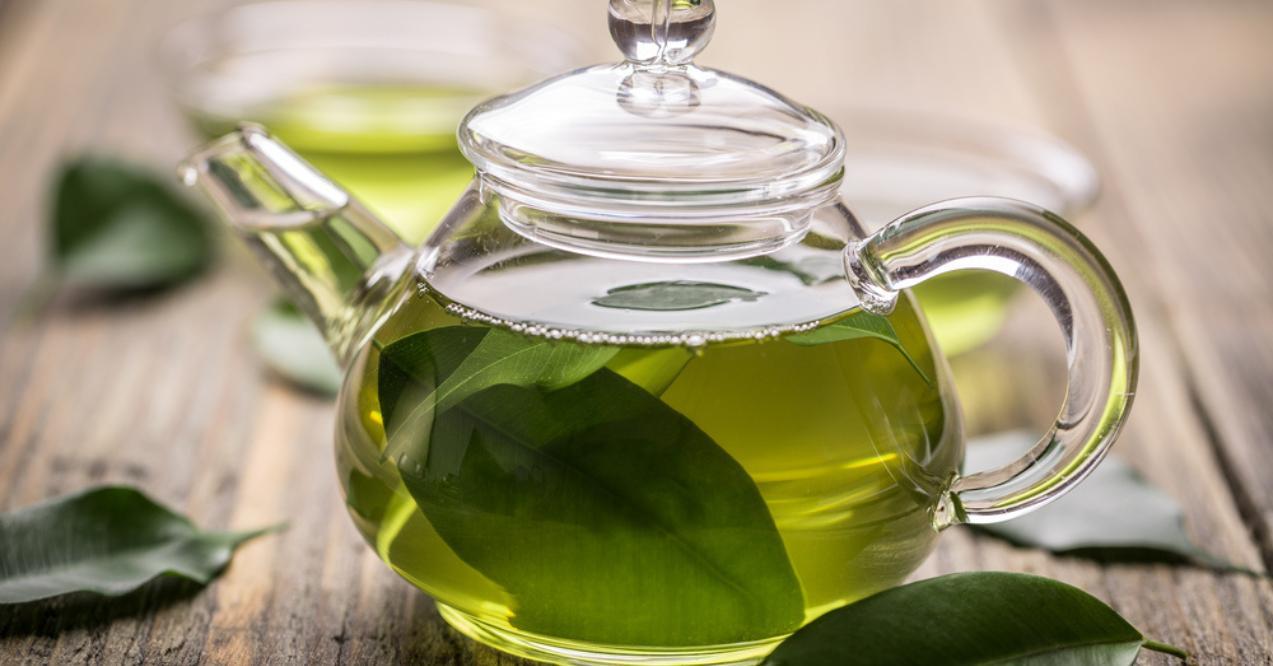 Clear glass teapot with green tea and leaves on a wooden table