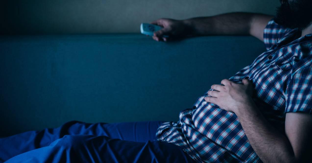 A person lying on a couch in a dark room, holding a remote, illustrating a sedentary lifestyle
