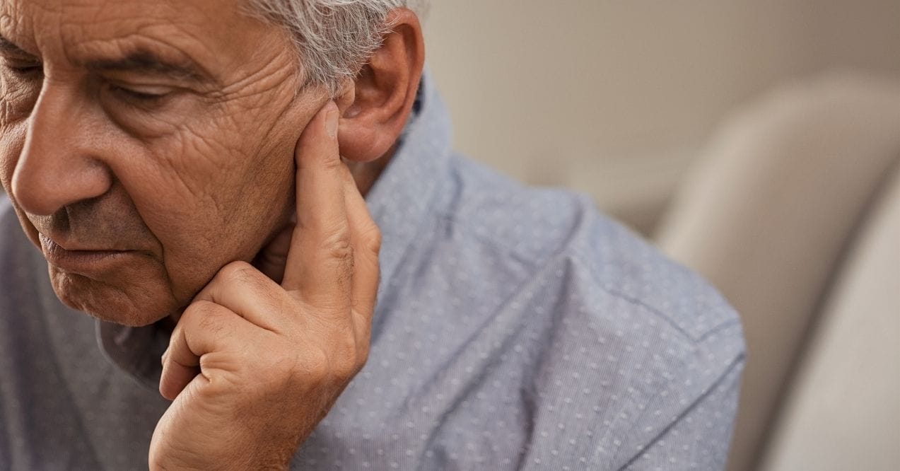 Side view of senior man with symptom of hearing loss. Mature man sitting on couch with fingers near ear suffering pain.