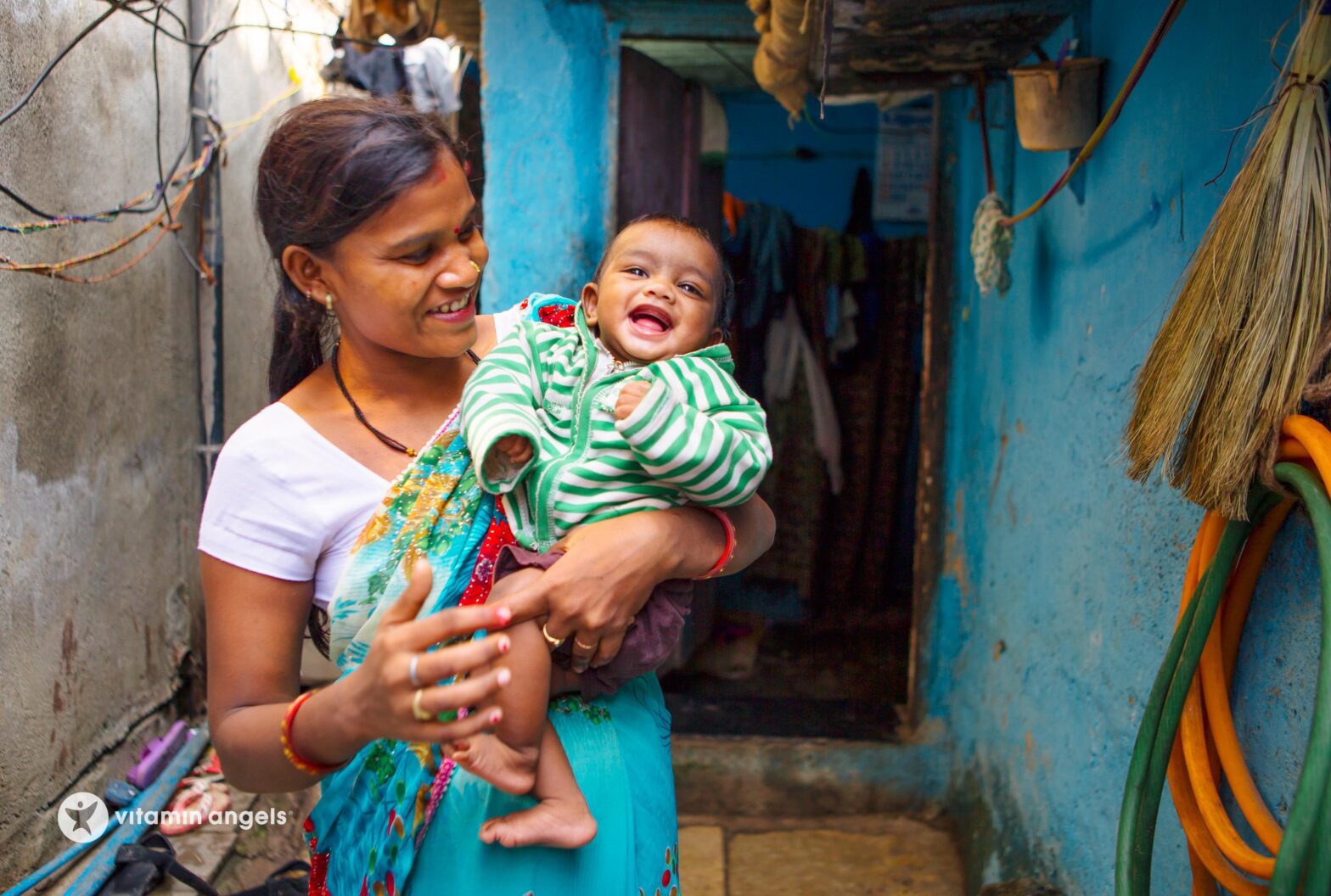 woman in a vibrant blue dress smiling as she holds a laughing baby, in a narrow blue alleyway