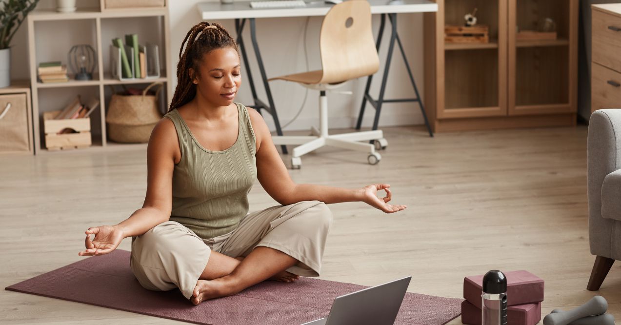 a woman sitting cross-legged on a yoga mat in a home setting, meditating with a laptop and yoga accessories around her