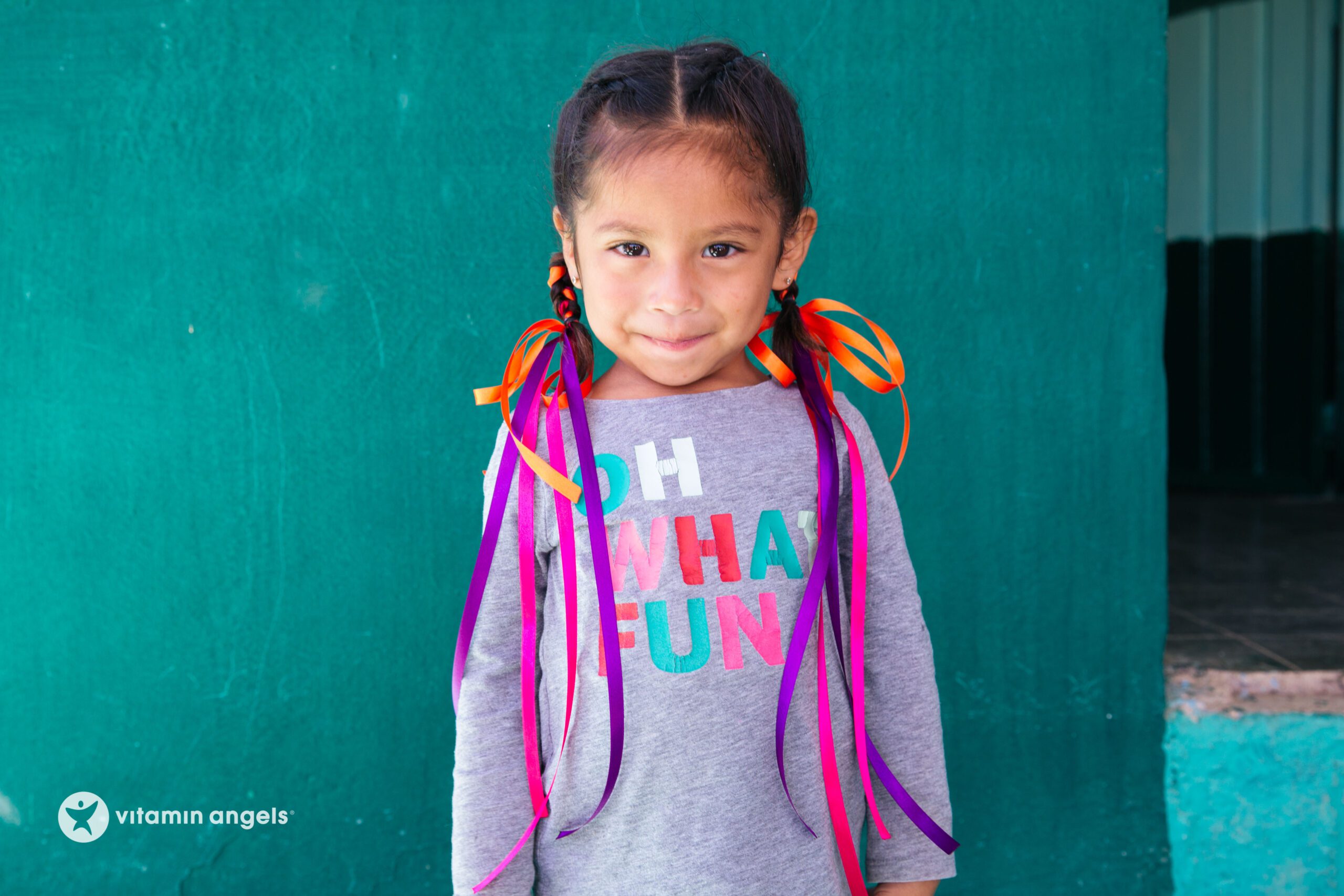 young girl with colorful ribbon braids, smiling in front of a turquoise wall, wearing a playful t-shirt