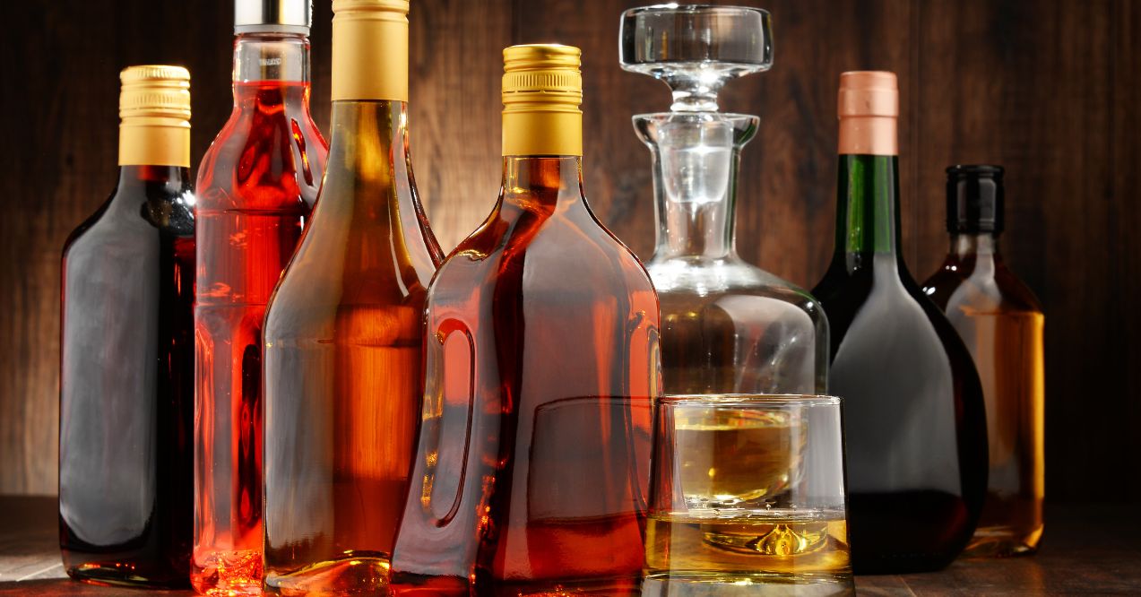 assorted bottles of alcohol, including wine and spirits, arranged on a wooden surface with a glass of whiskey in the foreground.