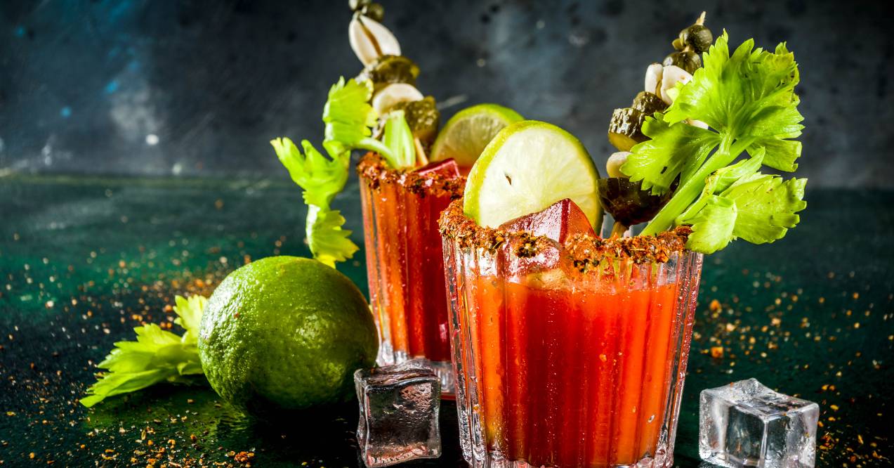 two glasses of Bloody Mary cocktails garnished with celery, pickles, and lime wedges, placed on a dark surface with ice cubes and lime.