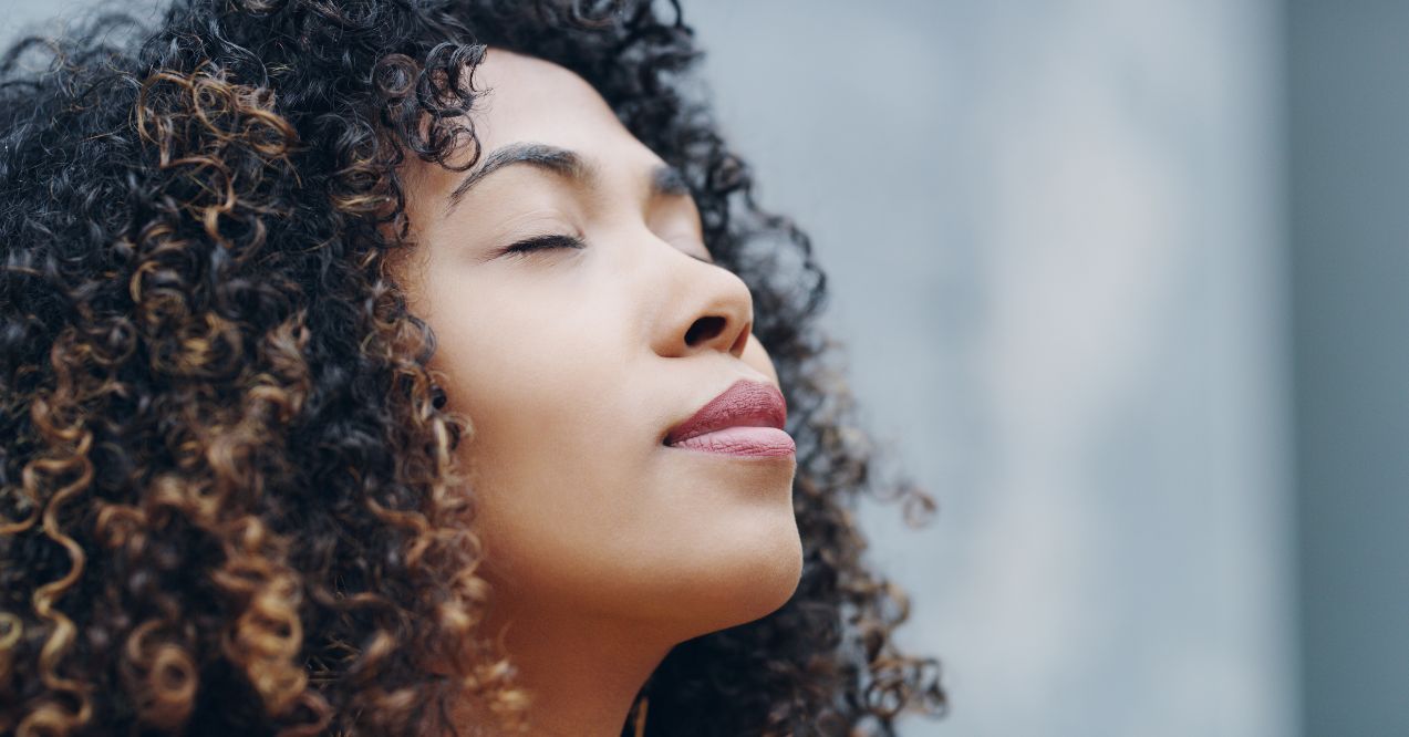 a woman with curly hair performing breathing techniques, eyes closed in a serene expression, set against a soft-focused urban background