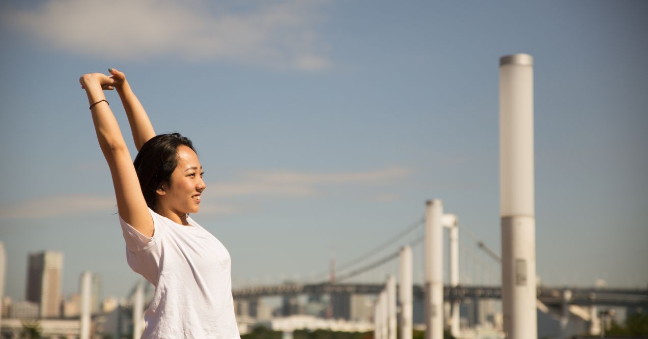 a woman standing outdoors stretching her arms upward, with a bright cityscape and a bridge in the background