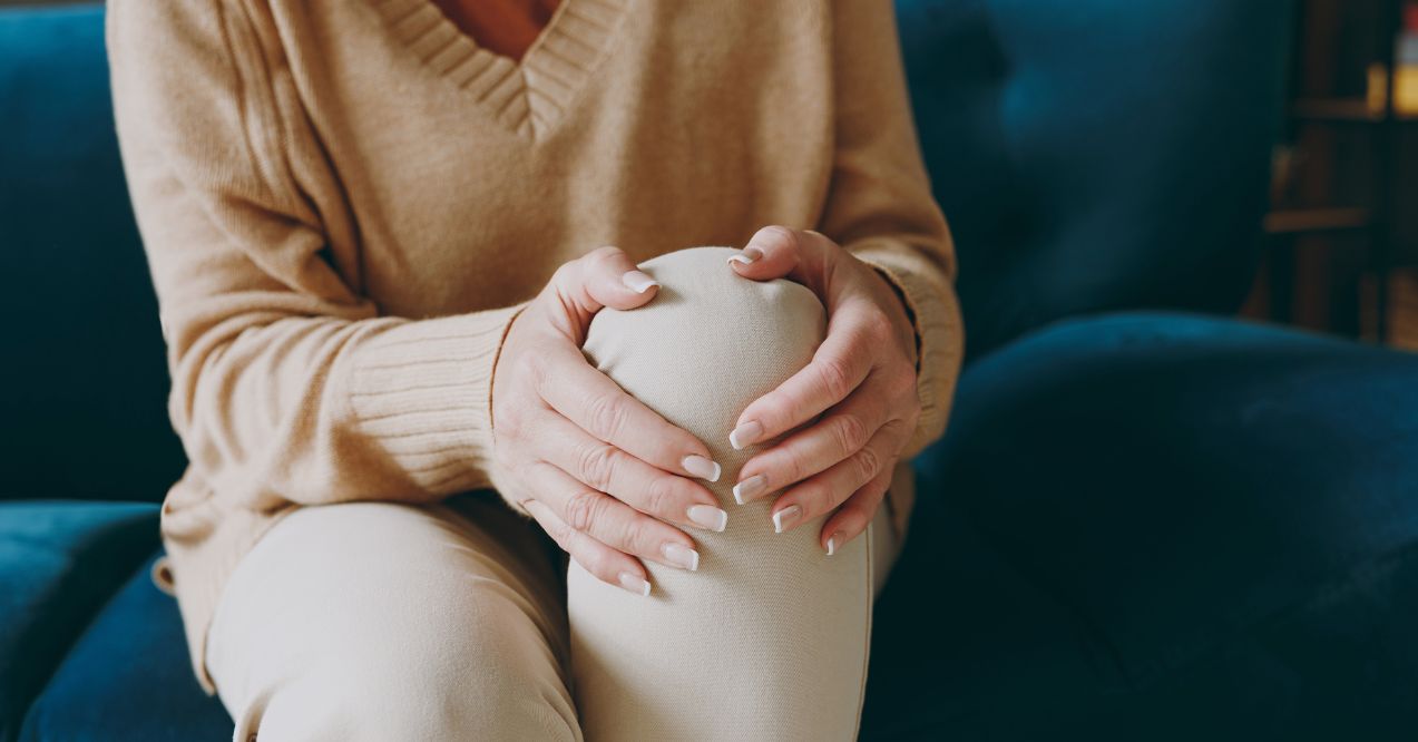 Cropped close up photo of pensioner woman wear casual clothes sits on blue sofa hold knee suffer from leg knee pain inflammation stay home flat spend free spare time in living room indoor grey wall.