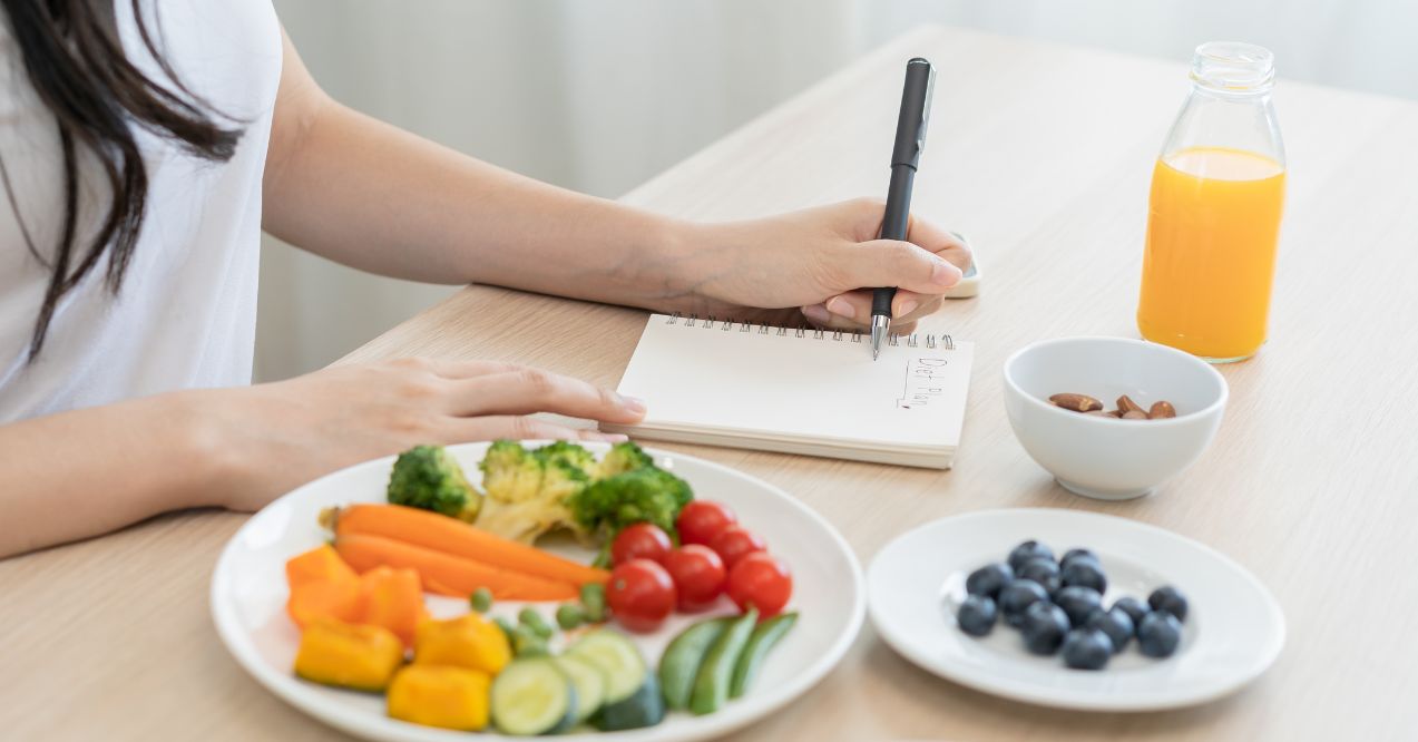 A woman writing down a keto diet plan to a notebook next to a plate of vegetables, blueberries and orange juice.