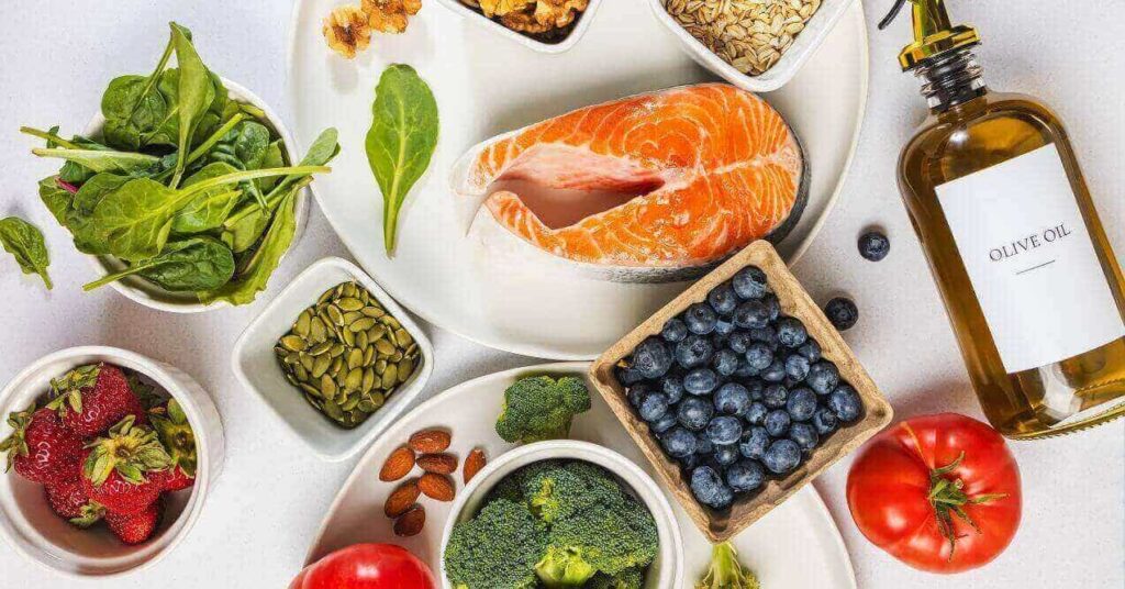 A colorful display of healthy, anti-inflammatory foods including salmon, blueberries, spinach, strawberries, pumpkin seeds, almonds, broccoli, walnuts, oats, and olive oil, arranged in small dishes and plates on a white surface.