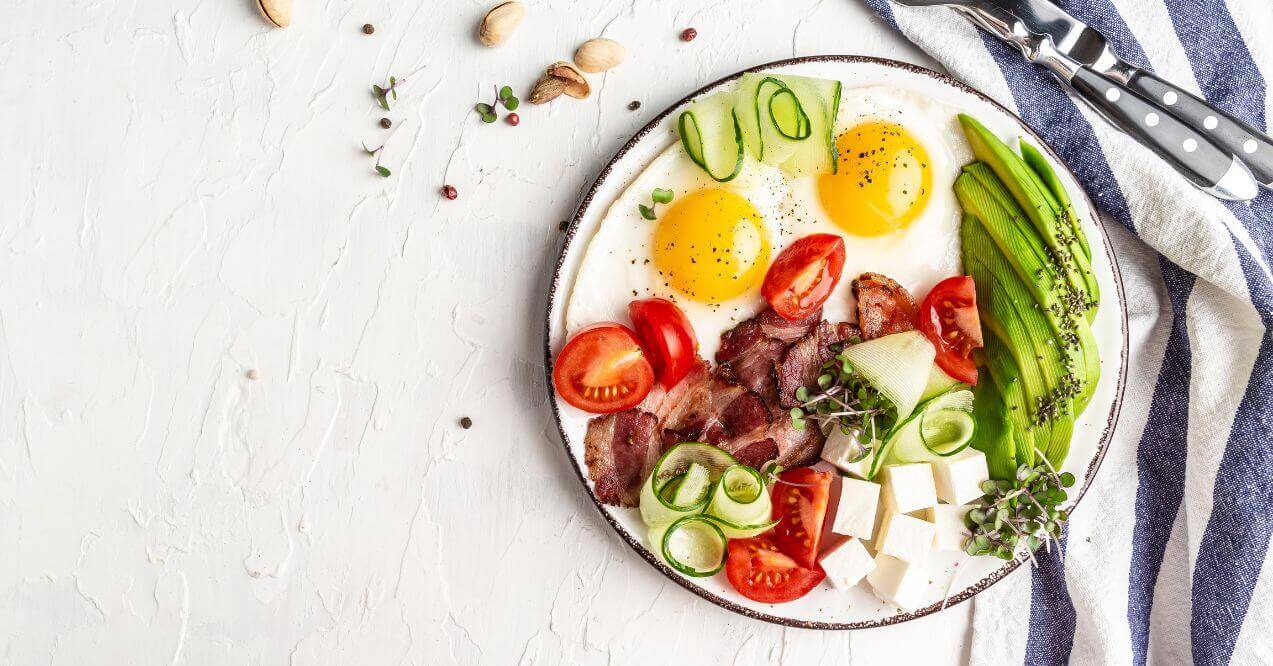 Keto-friendly breakfast plate with eggs, bacon, avocado, tomatoes, and cucumber slices, arranged on a white dish.