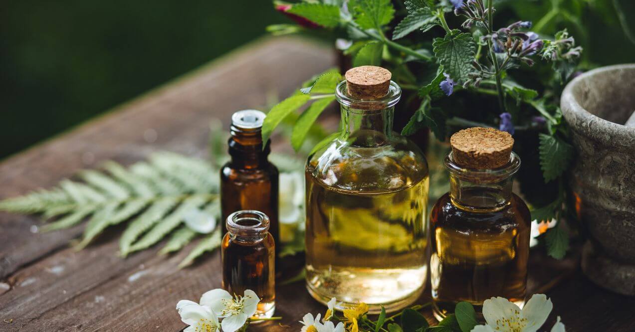 Various bottles of essential oils placed on a wooden table with fresh herbs and white flowers, used for toenail fungus treatment.