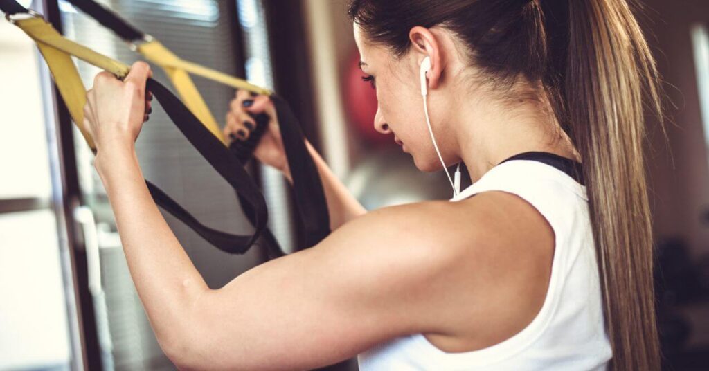 A woman with a ponytail wearing a white tank top, using suspension straps during a workout at the gym. She is focused on her strength training routine.