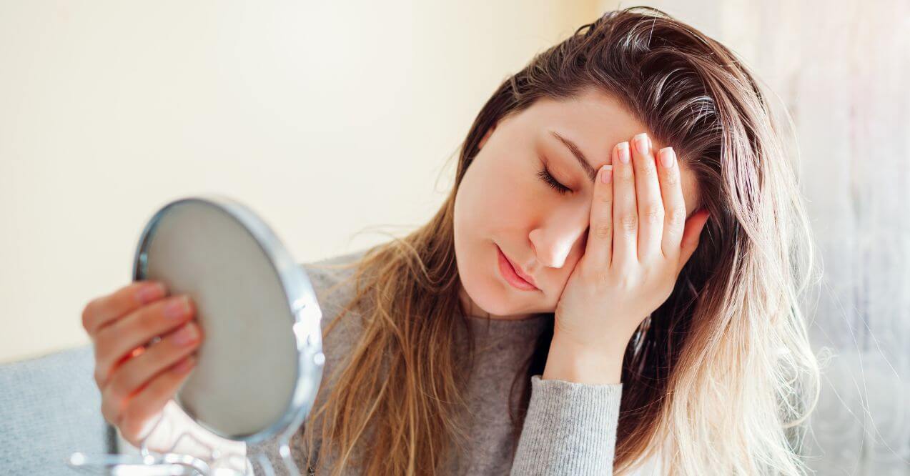 Young woman is holding her head after looking into a mirror because of her bad hormonal balance.