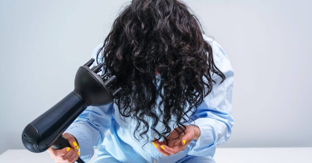 A dark curly haired woman holding her head down and drying her hair with a diffuser.