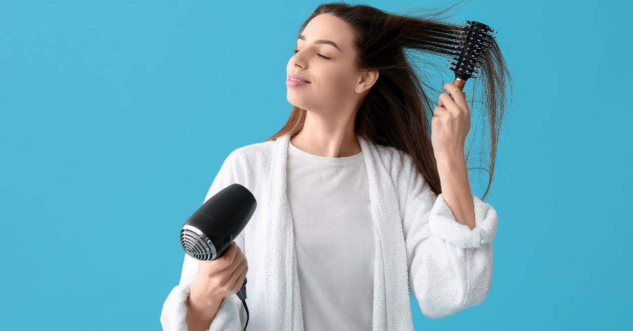 A young woman is drying her hair with a hairdryer and brushing it with the other hand.