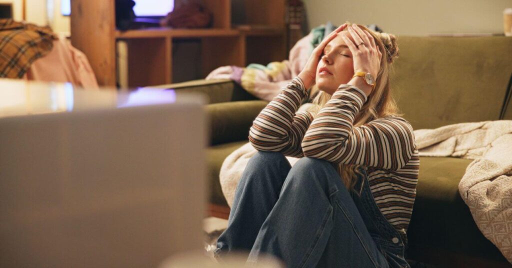 A woman sitting on the floor in a house and holding her head with both hands because of stress.