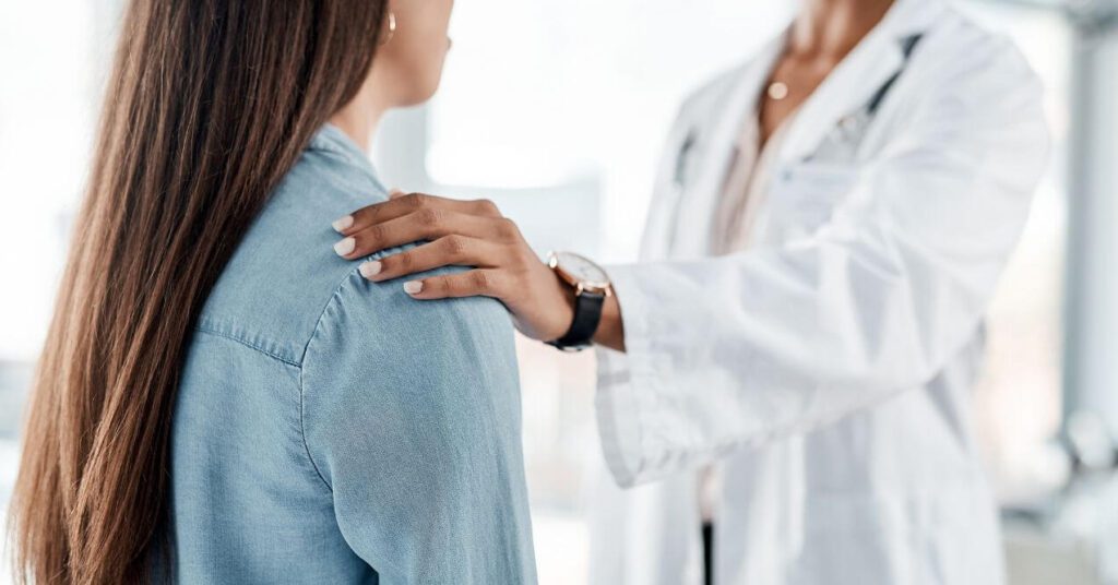 A woman is standing and looking at a doctor and the doctor is holding her shoulder because of bad news.