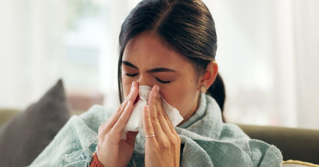 Woman, home and sinus with blowing nose on illness, flu and sick in living room on sofa. Female person, sneeze and couch in lounge with blanket for fever, virus and allergy with tissue for infection