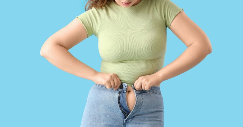 A woman is trying to button her pants but struggling because of weight gain.