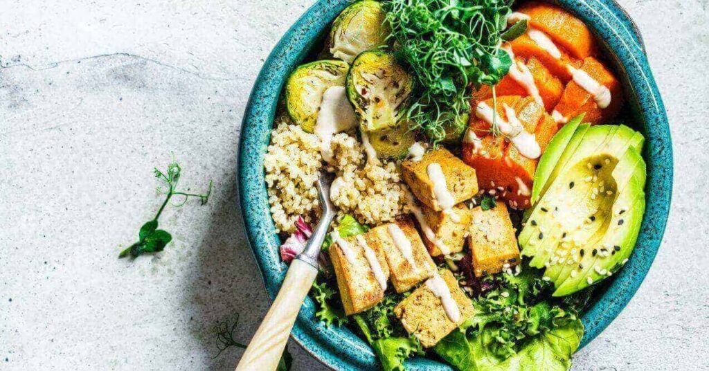 A vibrant bowl filled with nutritious ingredients including quinoa, roasted brussels sprouts, avocado slices, sweet potatoes, tofu, and leafy greens, drizzled with a creamy dressing.