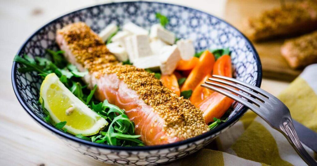 A bowl of fresh salmon salad featuring a sesame-crusted salmon fillet, arugula, lemon wedge, tofu cubes, and sliced vegetables such as carrots, offering a healthy and balanced meal.