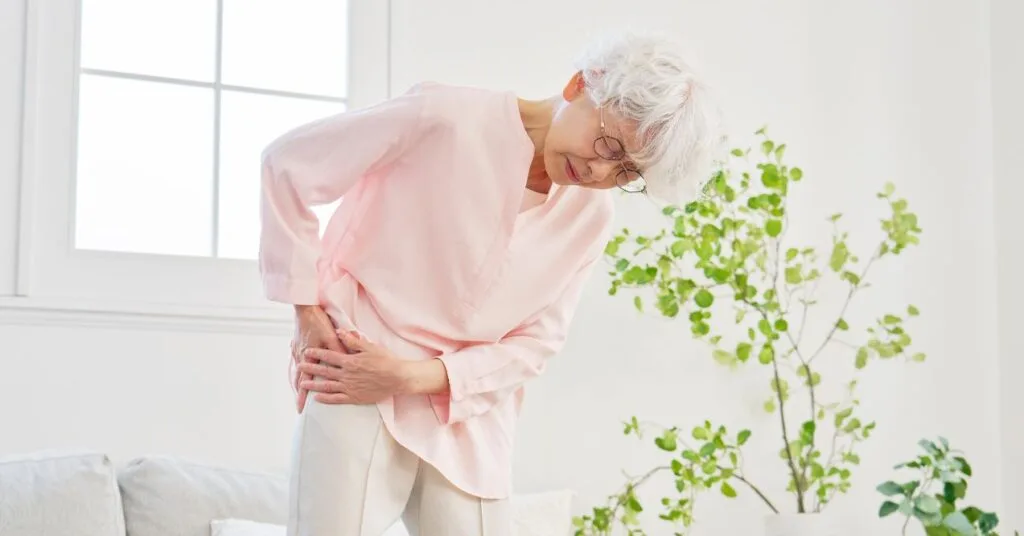 Asian senior woman with sciatica standing in the room and holding her hip because of the pain.