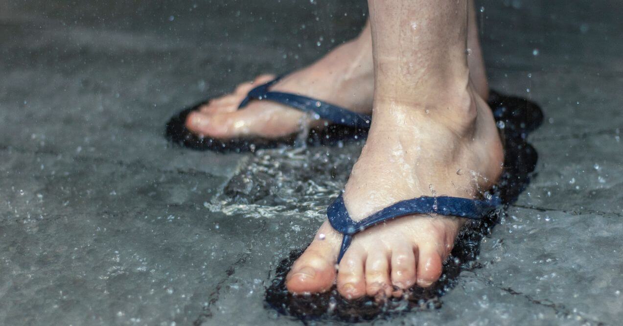 Feet wearing dark blue flip-flops in the shower as water splashes around.