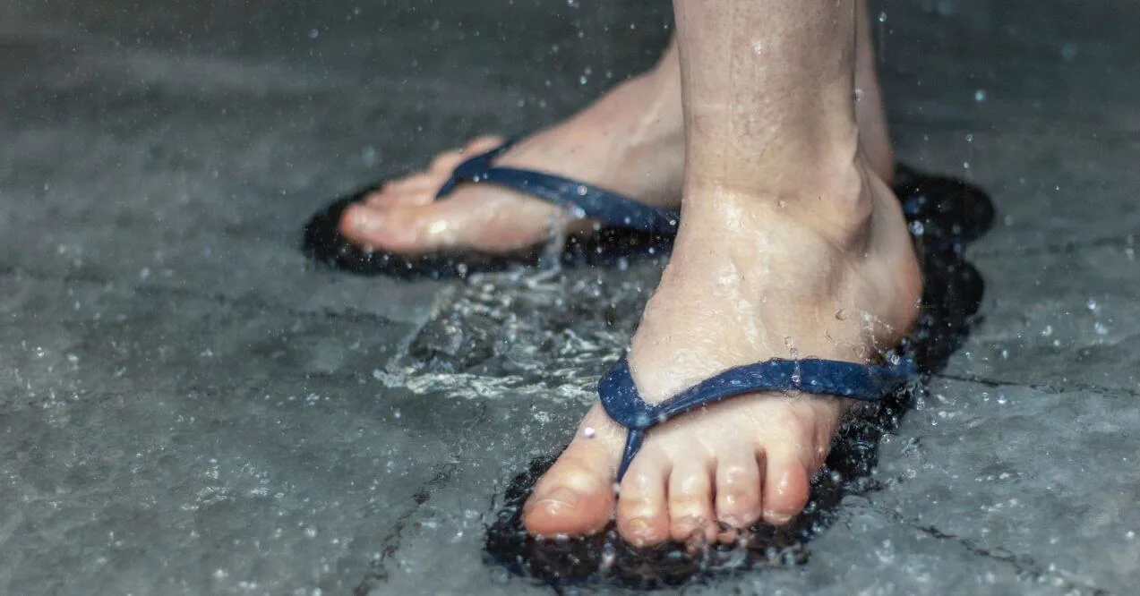 Feet wearing dark blue flip-flops in the shower as water splashes around.