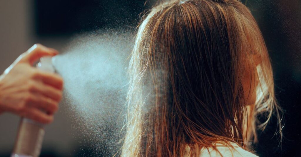 A young woman is facing sideways while the other person is spraying hairspray on her hair.