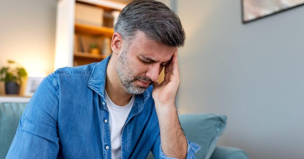 Man holding his head in pain, experiencing a headache caused by stress while sitting on a couch.