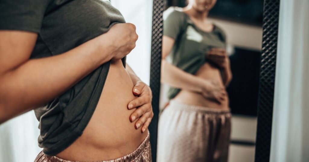 A woman is looking into the mirror and holding her bloated stomach because of digestion problems.