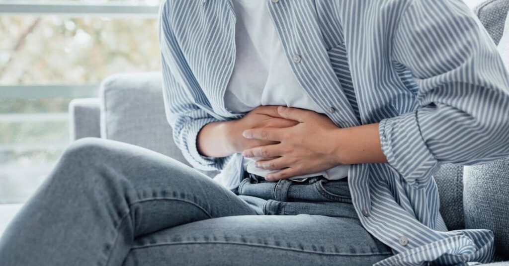 Person clutching their abdomen while sitting on a couch, showing signs of abdominal pain.