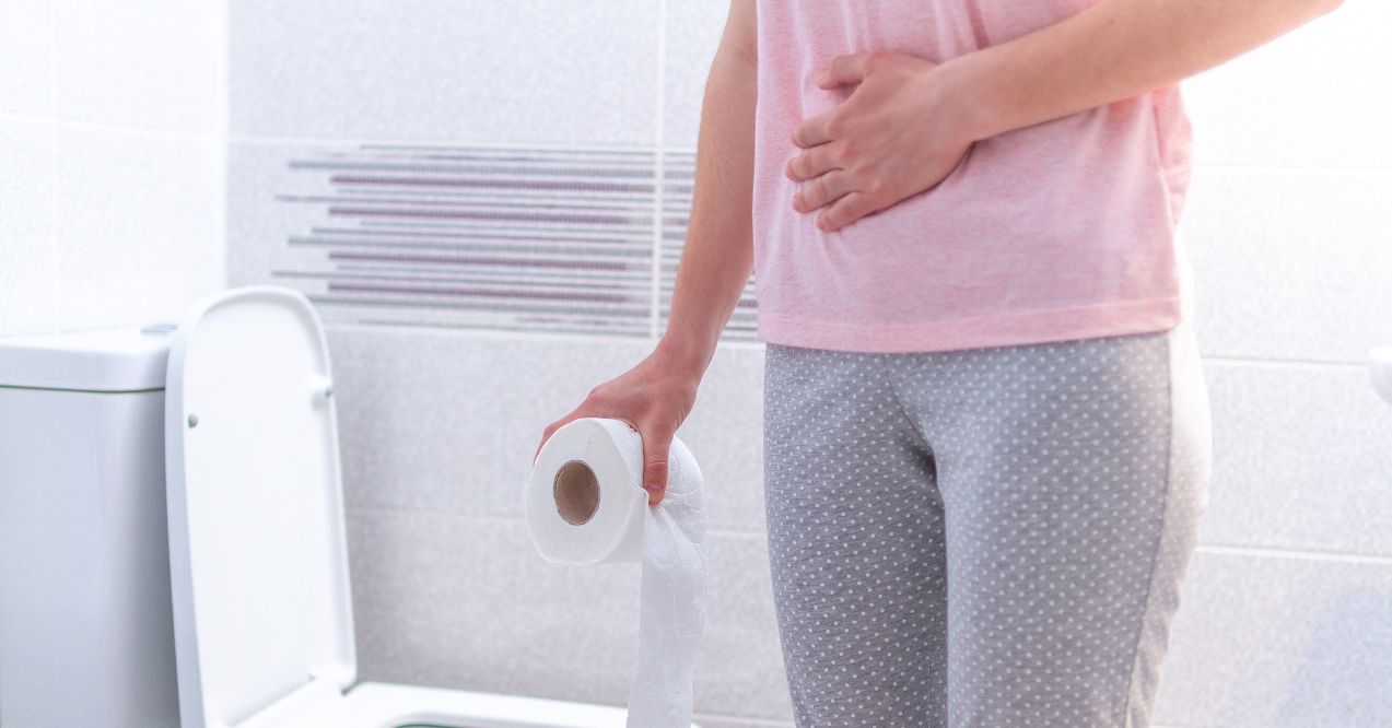 Person holding a roll of toilet paper while clutching their abdomen, standing near a toilet, indicating stomach discomfort.