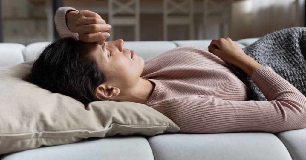 Woman lying on a couch with her hand on her forehead, displaying symptoms of fatigue and weakness.