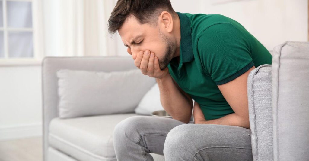 Man sitting on a couch, holding his stomach and covering his mouth, experiencing nausea.